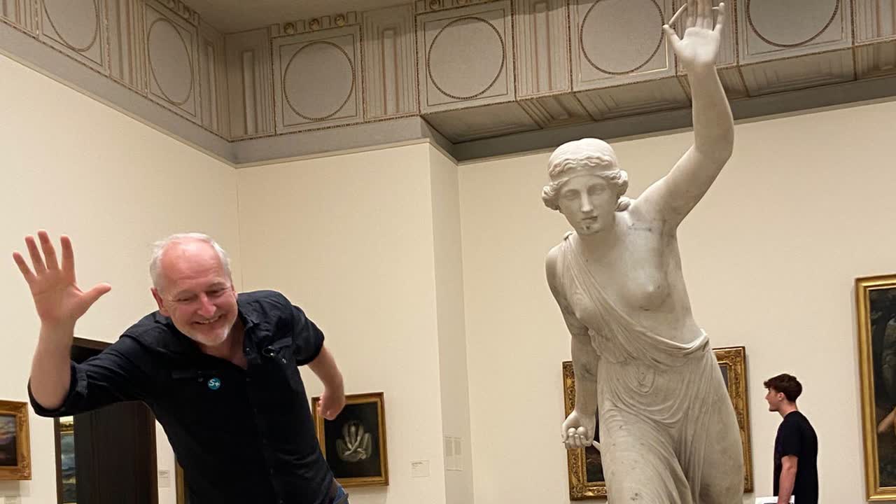 Een vrolijke man imiteert speels de pose van een wit marmeren beeld in een museumzaal