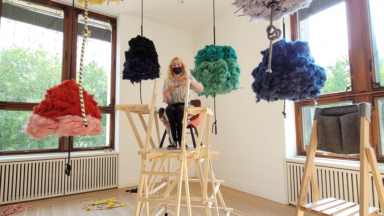 Een kunstenares werkt aan een installatie tussen kleurrijke, aan touwen hangende wolsculpturen in een lichte zaal
