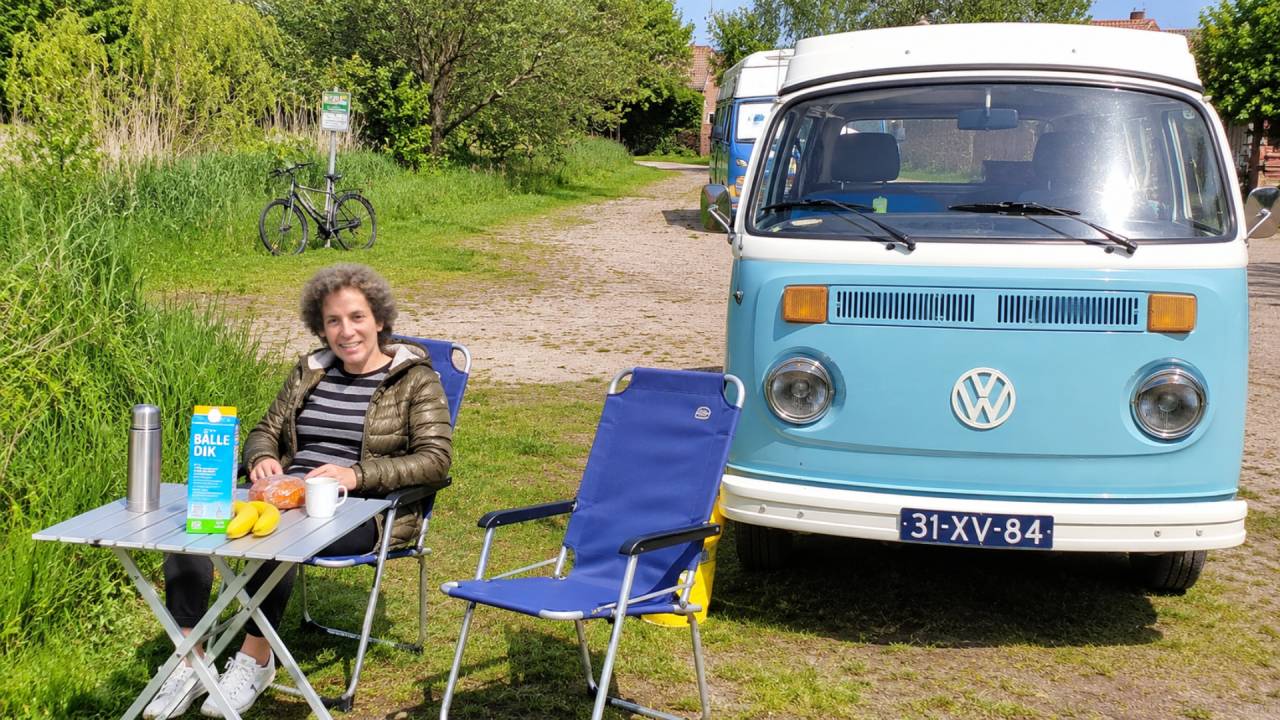 Een lachende vrouw ontbijt aan een campingtafel naast een lichtblauw Volkswagen-busje in het groen